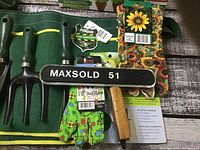 Green mesh tool apron with three small hand gardening tools (rake, fork, trowel) inserted, and pairs of colorful gloves arranged on a rustic wood surface.