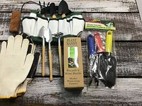 Garden tool bag with multiple pockets containing wooden-handled metal garden tools, white gloves with black grip, Plant Nanny watering device in box, and set of three colored handle garden tools in plastic packaging arranged on a wooden surface.