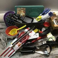 Overview photo showing small black raclette pans, kitchen utensils including strainer, tongs, ladle, spatulas, and cleaning brushes, green glass coaster set with floral prints, and metal spiral egg holders.