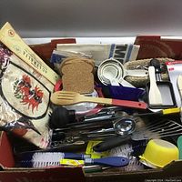 Photo showing a box containing various kitchen items such as white measuring cups, cork coasters, wooden spatula, metal utensils, can opener, cleaning brushes, funnel and other kitchen tools.