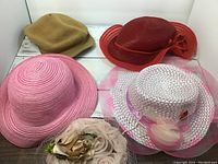 Photo showing four hats: tan soft fabric, red glossy with bow, pink wide-brimmed straw, white netted with pink feathers. All vintage style party hats.