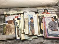 Several knitting pattern booklets and leaflets spread out on a wooden surface featuring different knitting designs for sweaters, throws, and men’s clothing.