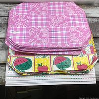 Stack of pink and fruit pattern vinyl placemats on rustic wooden surface.