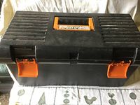Closed black plastic tool box with orange clasps on a patterned cloth surface.