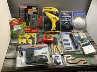 Full lot setup on table, showing tools in packaging and unpackaged items including stapler, hot glue gun, measuring tape, rotary cutter, caliper, driver set, dust mask, and adhesive hooks.
