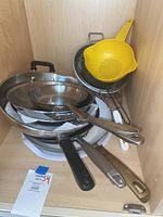 All Clad skillets stacked with strainers and colander in cabinet
