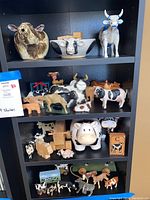 Wide shelf view showing assorted cow figurines and decor on four black shelves including large ceramic cow head planter and various small figurines and blocks.