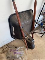 Craftsman wheelbarrow resting on a wall with visible black metal basin and wooden handles. Bag of stone base on floor.