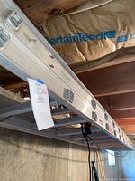 Long view of heavy duty aluminum extension ladder hung horizontally in basement space. Ladder is silver with metal rungs and locking hardware visible.