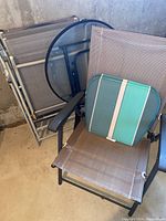 View of one folding outdoor chair with mesh fabric and armrests, two seat cushions on it, and folded chairs and table behind.