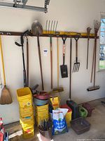 Wall of gardening tools hung from a horizontal support including shovels, rakes, pitchfork, broom and a yellow stack of plastic buckets beneath with watering cans and lawn repair bag nearby.