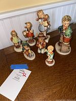 Photo showing all seven Hummel figurines arranged on a wooden table, depicting various children in different poses.