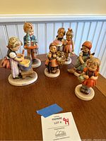Top view of 7 Hummel figurines on wooden table showing details of each figure's posture and clothing.