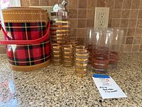 Complete lot showing plaid ice bucket, metal cocktail shaker, six gold-striped short glasses, and five tall brown-striped glasses on kitchen counter