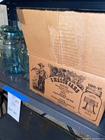 Cardboard box labeled 'Mason Jars' with clear glass jars and vintage bluish jars with metal clamp lids on a shelf.