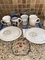 Lot contents: 7 decorative mugs in various designs, 2 ceramic lion relief wall plaques, and 1 ceramic lion face holder on a kitchen countertop.