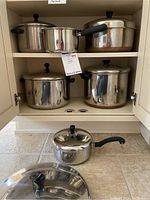 Cookware set of various stainless steel pots and lids arranged inside open kitchen cabinet and on floor.