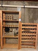 Two wood racks side by side in basement against concrete wall. One is a tall four-shelf unit with multiple items stored. Second is a large wine bottle rack with horizontal slots for bottles.