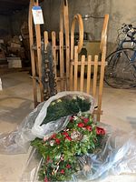 Two wooden sleds shown upright with vertical slat seats and curved back handles. Christmas wreaths with festive decorations are placed in front.