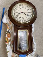 Full view of vintage wooden wall clock lying flat, showing the circular clock face with Roman numerals and date numbers around the edge, framed by dark wood case with glass lower door.