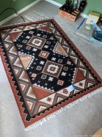 Full view of rectangular wool dhurrie style rug with earth toned geometric patterns on carpeted floor. Shows entire rug and fringe ends.