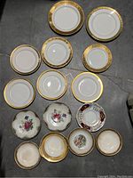 Top view showing all fifteen antique white china plates with various gold trim patterns and sizes, some with floral decorations and one with visible damage.