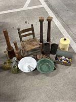 Wooden kitchen tools, small wooden chair with woven seat, metal graters, cast iron molds, enamelware bowls, old tins, and a box of assorted kitchen and sewing accessories arranged on concrete floor.