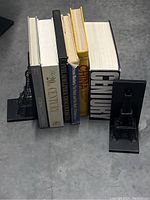 Photo shows six hardcover coffee table books upright between two black metal Eiffel Tower bookends on a concrete floor.