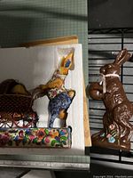 Large chocolate brown rabbit figurine holding egg, adorned with pink ribbon and blue flower, shown outside packaging for full visibility.