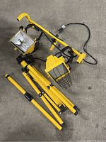 Yellow industrial work lamp light with protective cage and wired power cord on a concrete floor, tripod stand disassembled and numerous yellow metal pieces forming the stand.