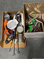 Wide overhead photo showing two cardboard boxes holding kitchen cutting tools, knives, shears, and miscellaneous small kitchen utensils and gadgets.