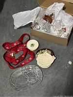 Photo of miscellaneous Christmas items including red suspenders, two decorative plates, a clear glass tray, and a box of Christmas ornaments wrapped in white tissue paper.