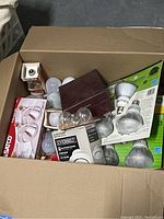 Top-down view of open box showing mixed assortment of packaged and loose lightbulbs including Satco and Everbilt brands.