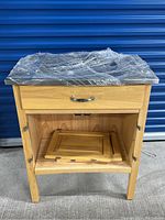 Front view showing wooden cabinet with drawer and two detached doors placed inside cabinet interior. Metal top covered with clear plastic wrap.