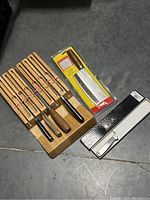 Photo showing multiple kitchen cutting tools including sharpening steels, a knife, and sharpening stones in packaging along with a wooden holder.