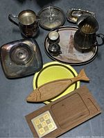 Various silverplate serving pieces including pitchers, covered dish, trays, and small cups alongside wooden and metal trays on a dark surface.