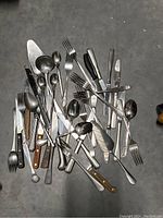 Pile of assorted flatware including forks, knives, spoons with various handle materials including wood and metal, showing usage wear and tarnish.