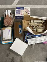 Top view photo showing box of typewriter ribbons, two computer keyboards, reels of dot matrix printer paper, electrical cords, and miscellaneous office items on a carpeted floor.