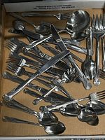 Photo showing the complete collection of flatware pieces arranged inside a cardboard box, including forks, knives, spoons, tablespoons, teaspoons, and serving ladle, all featuring a retro style handle design.