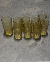 Set of 9 amber-colored drinking glasses arranged in two rows on a concrete floor, showing varying heights and textured wood grain design throughout the glass surfaces.