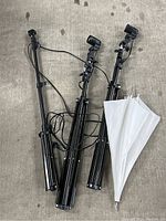 Two black adjustable tripods each with mounted spotlight photography lights and white photography umbrellas folded and attached.