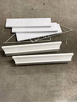 White wall shelves shown stacked on carpet floor with four flat shelves, two decorative-edged shelves, and two metal brackets.