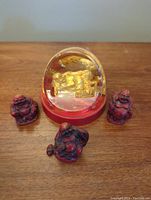 Three small red Buddha figurines and one large golden Buddha pencil and paperclip holder under a clear dome, all on a wooden surface.