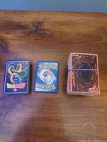 Three stacks of trading cards: Yu-Gi-Oh (heavily used, rubber banded), Pokémon cards with classic back design, and a Dragon Ball Z card box featuring Shenron.