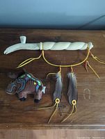 Lot showing both items together on wooden surface: the antler peace pipe with carved twisted handle and three hanging feathers, and the plush moose with indigenous design embroidery.