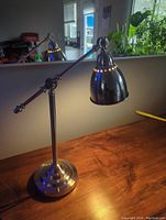 Photo showing the lamp turned on, highlighting polished stainless steel surface and weighted base on wooden surface.