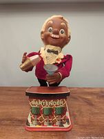 Front view of bartender toy showing head, face, velvet jacket with bow tie, cocktail shaker, martini glass, and metal base with 'BARTENDER' text.