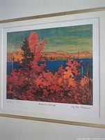 Close-up image of the framed print showing the autumn scene with vibrant red and orange foliage, blue water body, and hills in the background, with handwritten numbering and title beneath the print.