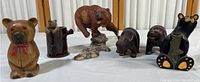 Seven bear figurines displayed on a white surface, including wooden bears, a porcelain/ceramic bear on a rocky base, and painted wooden bears