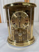 Front view of gold-tone Benchmark clock showing Roman numeral dial, gold finish, and four orb pendulum balls inside glass case.